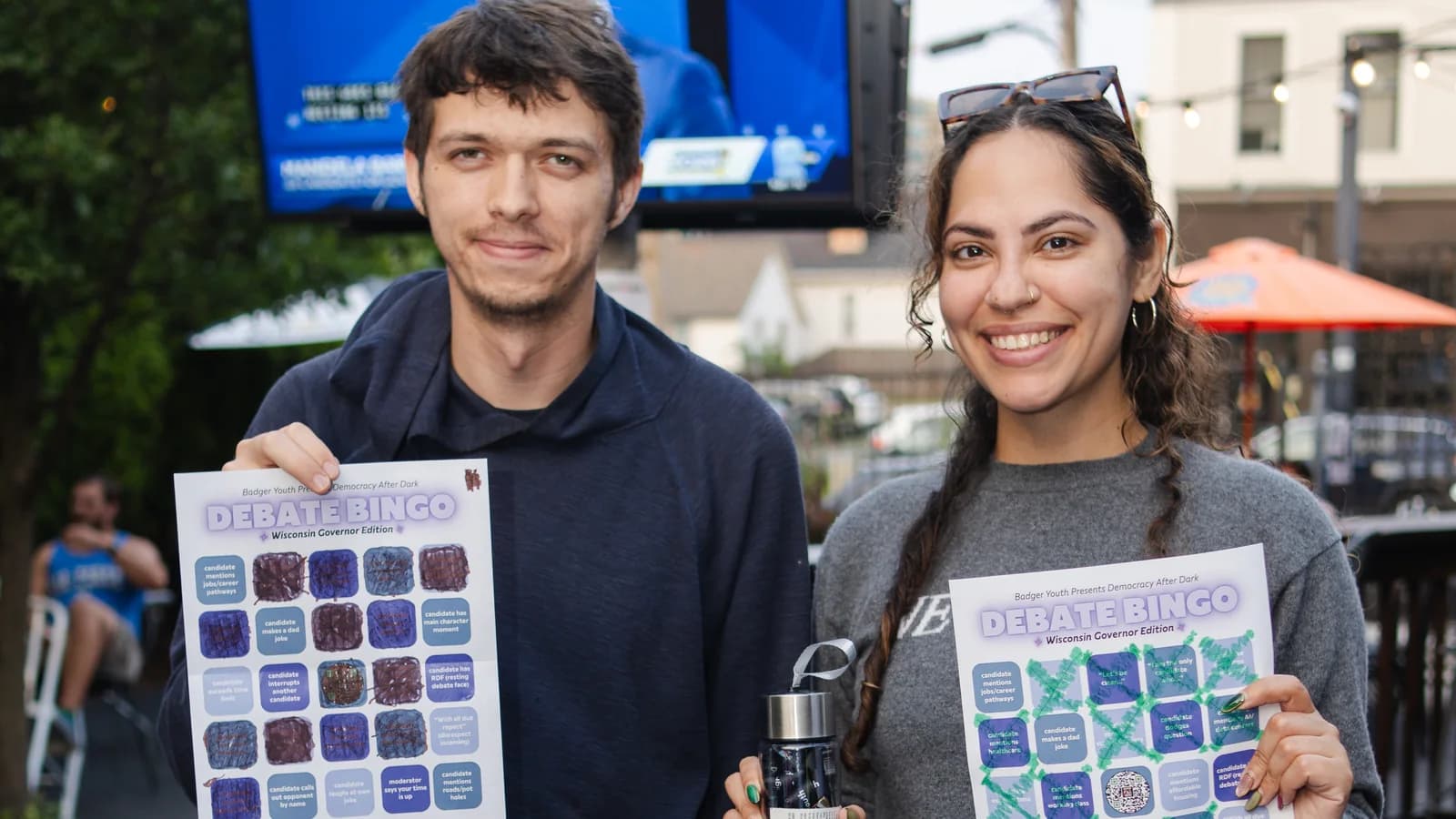 Two young Wisconsin organizers hold completed Debate Bingo cards at an outdoor gathering.